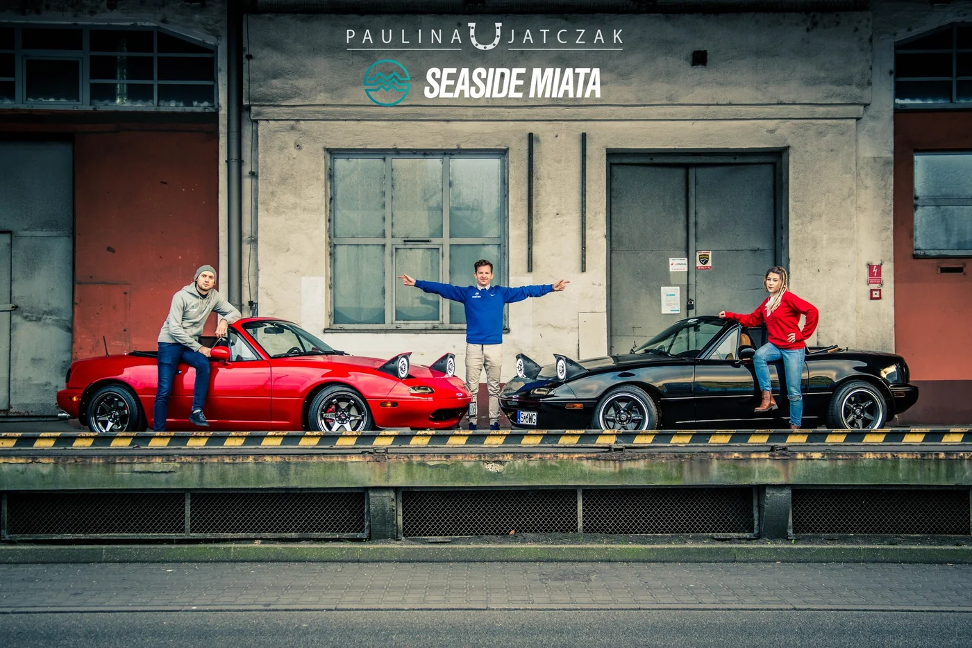 Czerwona i czarna Mazda MX-5 z trzema osobami przed industrialnym budynkiem